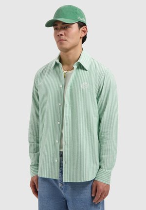 Jonge man die een groen-wit gestreept overhemd met knopen, lichtblauwe spijkerbroek, een groene pet en gouden kettingen draagt, tegen een effen achtergrond.