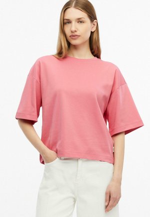 Jeune femme portant une chemise rose ample à manches courtes et un pantalon blanc, debout avec une main dans la poche sur fond blanc.