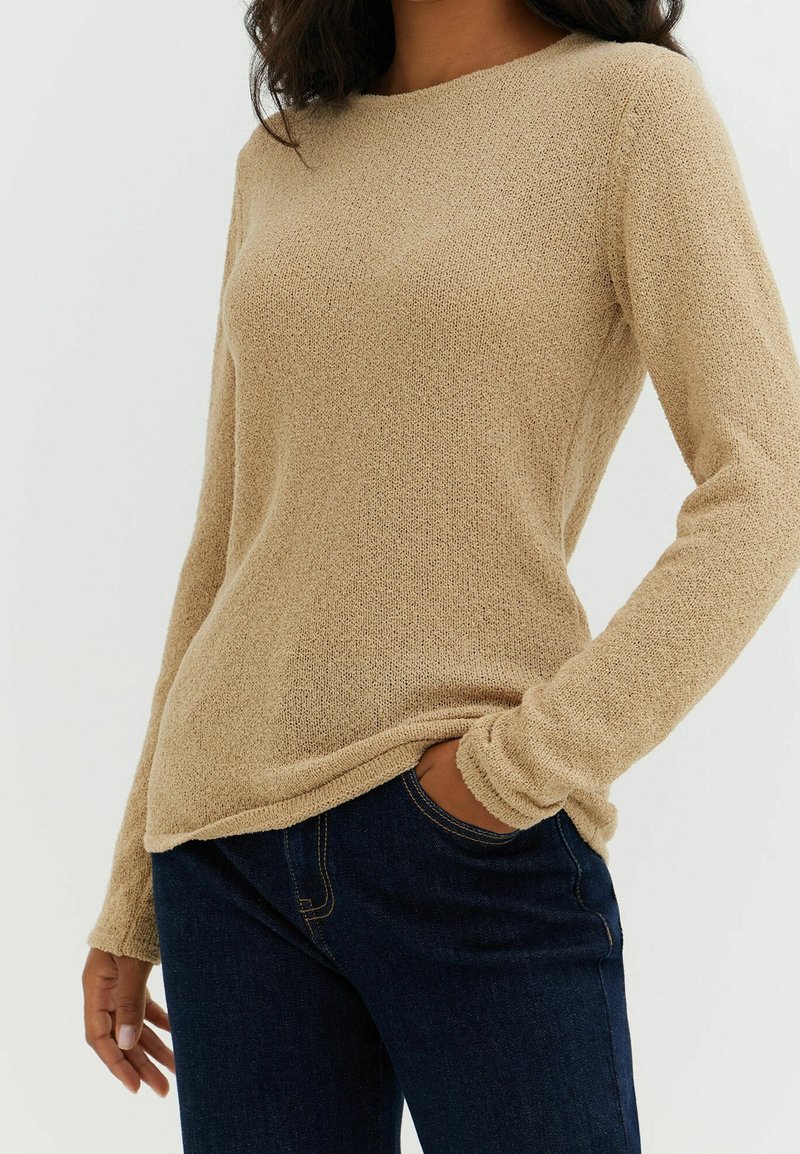 Femme portant un pull beige texturé à manches longues et un jean bleu foncé, une main dans la poche, sur un fond uni.