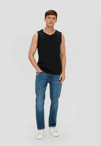 Black sleeveless shirt with a v-neck, worn with blue jeans. Model stands with one hand in pocket, wearing white sneakers. Plain gray background.