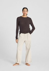 Top fitted a maniche lunghe a costine marroni abbinato a pantaloni cargo a gamba larga color crema con tasche laterali, in piedi su un pavimento nudo contro uno sfondo chiaro.