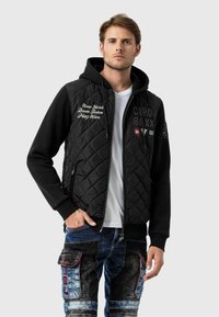 Schwarze gesteppte Jacke mit Kapuze, versehen mit gesticktem Text und Aufnähern. Kombiniert mit blauen Denim-Cargo-Hosen mit mehreren Taschen.