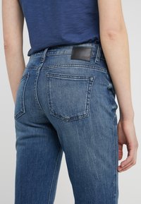 Modré džínsy s polovičním pasem, pěti kapsami a černou koženou nášivkou na zadním pasu. Vybledlá textura a rovný střih nohavic.