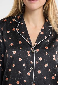 Czarna satynowa koszula do piżamy z białą lamówką, ozdobiona małymi różowymi kwiatowymi wzorami i kropeczkami. Zapięcie na guziki z przodu, krótkie rękawy.