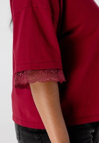 Een arm in een kortmouw bordeaux shirt met kant aan de mouwranden, gecombineerd met donkergrijze broek, tegen een effen lichte achtergrond.