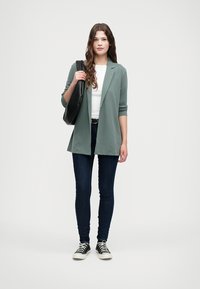 Lys grønn jakke, hvit topp og mørke jeans. Modellen holder en svart veske og har på seg svart og hvite sneakers. Enkelt design, casual passform.