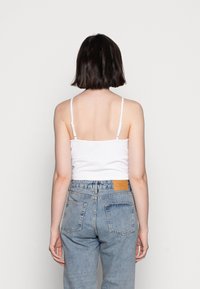 Top de alças finas, justo ao corpo, na cor branca, combinado com jeans de cintura alta em denim azul claro, com bolsos traseiros e um rótulo de couro bege.