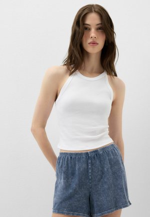 Ung kvinde med mellembrunt hår bærer en hvid ærmeløs ribbet top og blå elastiske shorts med taljebånd og står foran en ensartet lys baggrund.