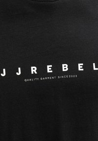 Μαύρο ύφασμα με λευκό κείμενο που γράφει "JJ REBEL" και μικρότερο κείμενο "QUALITY GARMENT SINCE 2023" τυπωμένο από κάτω.