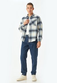Camisa de franela de cuadros en azul, blanco y beige, con un bolsillo en el pecho, combinada con jeans de mezclilla azul oscuro y zapatillas beige.