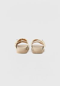 Kaksi takanäkymä sandaalia, joissa on teksturoitu kerma strap, beige muotoiltu pohja ja vaaleanruskea kumipohja. Yksinkertainen ja avoin malli.