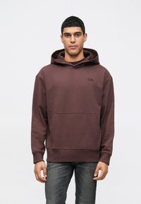 Άνδρας φορώντας σκούρο μωβ φούτερ με τσέπη καγκουρό και σκούρα γκρι τζιν, στέκεται μπροστά σε μια απλή ανοιχτόχρωμη φόντο.