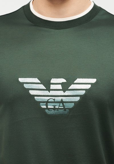 Άνδρας που φοράει σκούρο πράσινο πουκάμισο με λαιμόκοψη και λευκό και ανοιχτό μπλε στυλιζαρισμένο λογότυπο αετού και γράμματα "GA" στο στήθος.