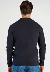 Mörkblå ribbad stickad tröja med rundad hals och långa ärmar; har texturerad väv och en sidetikett, kombinerad med blå denimjeans.