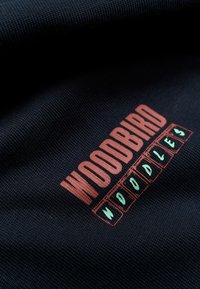 Σκούρο ύφασμα με τη λέξη «WOODBIRD» σε μεγάλα πορτοκαλί γράμματα πάνω από μικρότερα πράσινα γράμματα που σχηματίζουν τη λέξη «WOODLIES» μέσα σε περιγράμματα κουτιών.