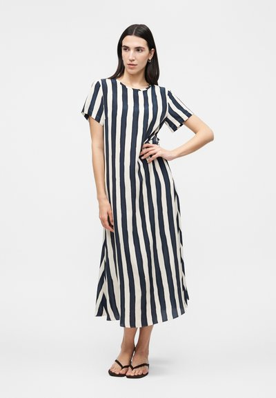 Γυναίκα στέκεται με το χέρι στη μέση, φορώντας μαύρο-λευκό midi φόρεμα με κάθετες ρίγες και μαύρες σαγιονάρες σε λευκό φόντο.