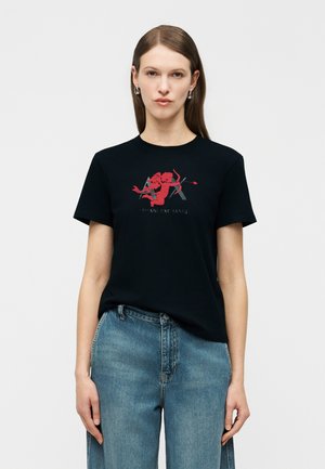 Žena v čiernom tričku s červenou grafikou Kúpeľa a nápisom "Armani Exchange", kombinovaná s modrými džínami s vysokým pásom, na jednotnom pozadí.