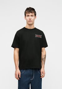 Zwart katoenen t-shirt met een grafisch ontwerp in het rood en wit op de linkerkant van de borst. Korte mouwen en een geribde ronde hals.
