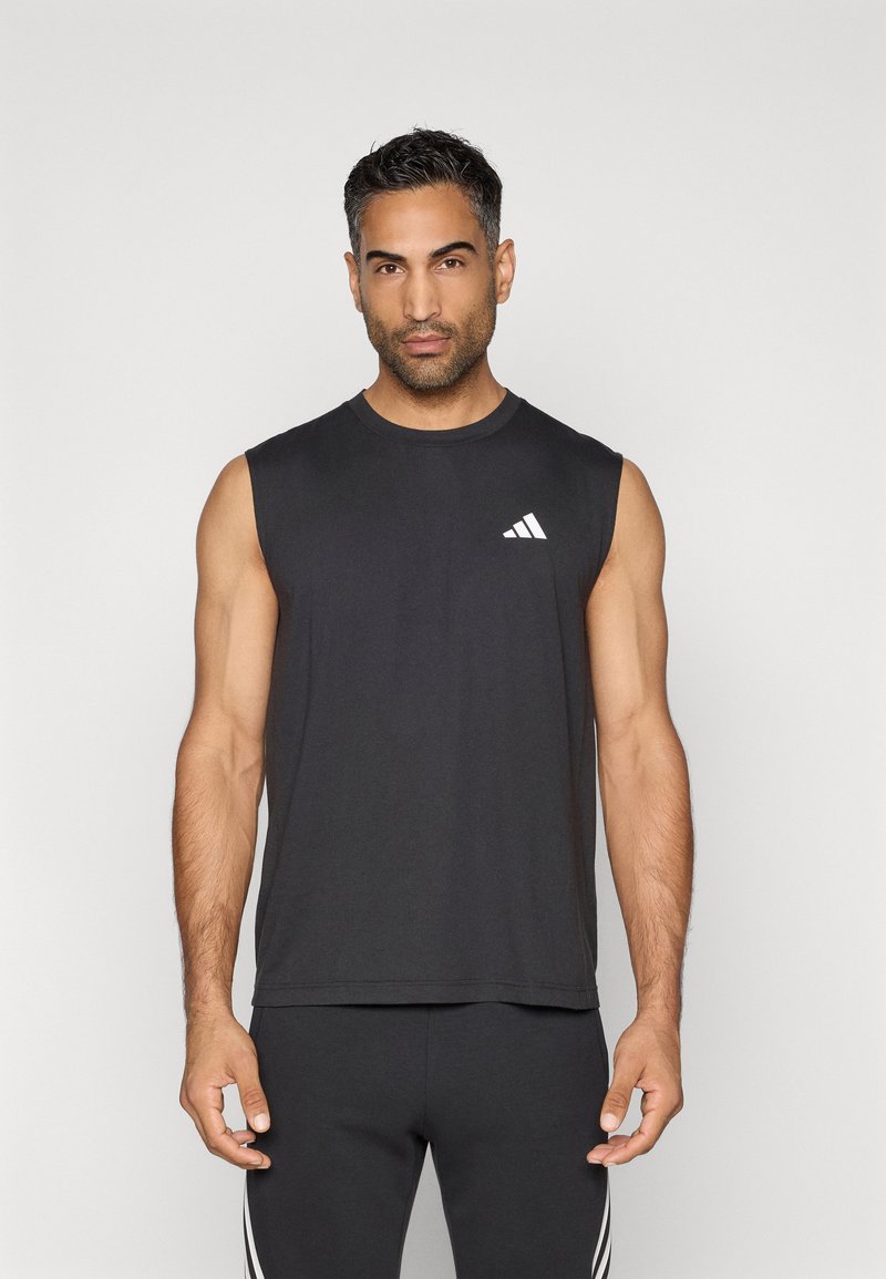 Férfi fekete ujj nélküli Adidas pólóban és fekete nadrágban, fehér csíkkal, egy sima világos háttér előtt áll.