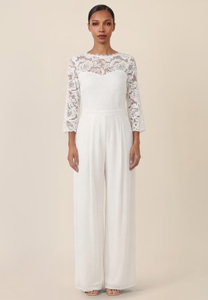 Frau trägt einen weißen Spitzen-Jumpsuit mit Dreiviertelärmeln und weit geschnittenen Hosenbeinen, steht vor einem beigefarbenen Hintergrund.