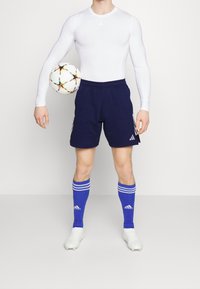 Camisa de compresión blanca de manga larga, pantalones cortos azul marino con rayas, calcetas de fútbol azules y botas de fútbol blancas, sosteniendo un balón de fútbol multicolor.