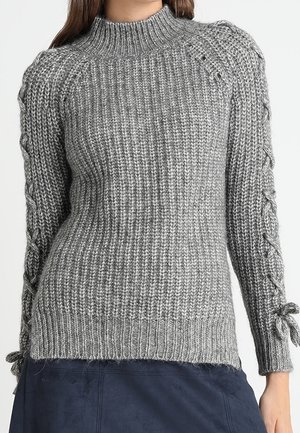 Pull en maille côtelée grise avec col montant et détails lacés sur les manches longues, porté avec une jupe sombre.