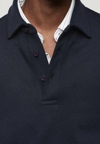 Polo shirt blu navy con design a colletto, caratterizzato da tre bottoni viola scuro e un tessuto texturizzato. Sotto, è visibile una camicia a righe bianche.
