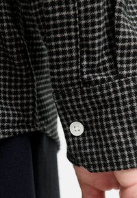Camisa a cuadros negra y gris con un tejido texturizado, que presenta un botón blanco en el puño y un dobladillo recto.