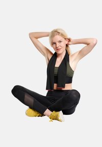 Athletic outfit featuring a black scarf, olive green sports bra, and black leggings with mesh accents. Model wears yellow sneakers.
