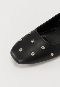 Melna plata kurpe ar kvadrātveida purngalu, teksturēta āda, rotāta ar sudraba kniedēm un rhinestones, kas sakārtoti rakstā pa virsmu.