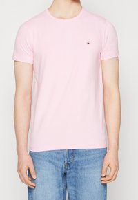 Mann in einem hellrosa T-Shirt mit kurzen Ärmeln und kleinem Logo auf der Brust, trägt eine hellblaue Jeans und steht vor einem schlichten Hintergrund.