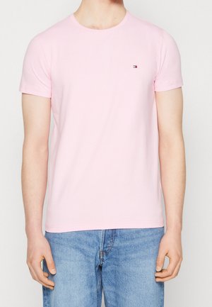 Man in een lichtroze T-shirt met korte mouwen en een klein logo op de borst, in combinatie met lichtblauwe jeans, staand tegen een effen achtergrond.