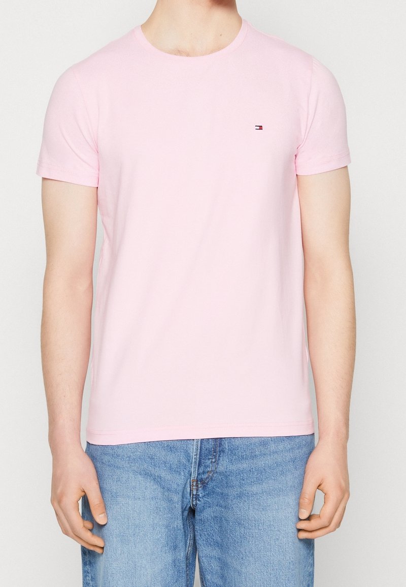 Mann in einem hellrosa T-Shirt mit kurzen Ärmeln und kleinem Logo auf der Brust, trägt eine hellblaue Jeans und steht vor einem schlichten Hintergrund.