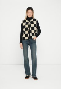 Gilet a scacchi nero e crema con bottoni su un dolcevita nero, abbinato a jeans blu svasati e scarpe con plateau nere.