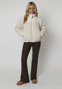 Mujer de pie con las manos en los bolsillos, vistiendo una chaqueta de forro polar crema, pantalones marrón oscuro y zapatos sin cordones color beige sobre fondo blanco.