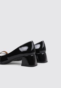 Zapatos de charol negro con tacón cuadrado, textura suave, punta redonda y detalles de correa en el tobillo.