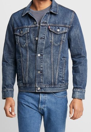 Mand iført en knappet blå denimjakke over en grå skjorte, kombineret med blå jeans, foran en ensfarvet lys baggrund.