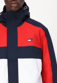 Rød, marineblå og hvit fargeblokkerende jakke med høy krage, framtidig glidelås og liten "Tommy Jeans"-logo på brystet. Materialet ser glatt ut.
