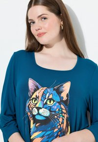 Camiseta de manga larga en color azul verdoso que presenta un gráfico colorido de la cara de un gato con ojos verdes. La tela tiene una textura suave y un escote redondo.
