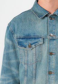 Light blue denim jacket featuring a classic collar, two chest pockets, silver buttons, and subtle fading patterns on the fabric.