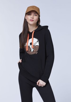 Schwarzer Hoodie mit orangefarbenen Kordeln, ausgestattet mit einem Grafikdruck eines Vogels und einem Sonnenuntergang. Das Modell trägt schwarze Hosen und eine beige Mütze.