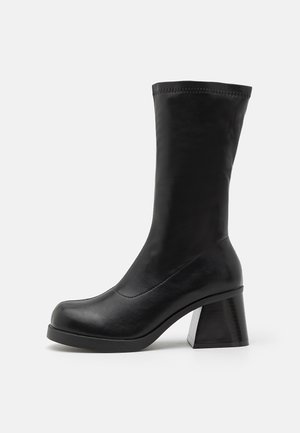 Svarte lærankelstøvler med en strømlinjeformet fasong, bred blokkhelt, og sydd detaljer langs toppkanten.