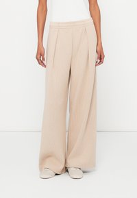 Béžové nohavice s širokými nohavicami s textúrovaným povrchom, skladaným predným dielom a elastickým pásom, skombinované so svetlou obuvou.