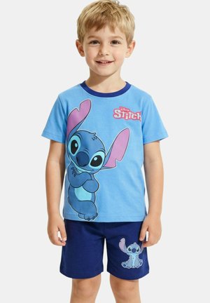 Jeune garçon portant un t-shirt bleu à thème Stitch et un short bleu marine, souriant et posant devant un fond uni.