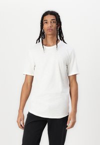 Weißes T-Shirt mit kurzen Ärmeln und abgerundetem Saum aus strukturiertem Stoff, kombiniert mit schwarzen Jeans. Modell steht vor einem schlichten Hintergrund.