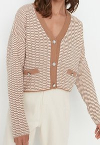 Háčkovaný cardigan v béžové a krémové barvě s výstřihem do V, předním zapínáním na knoflíky, dvěma kapsami a dekorativními knoflíky ve tvaru perel. Měkká pletená textura.