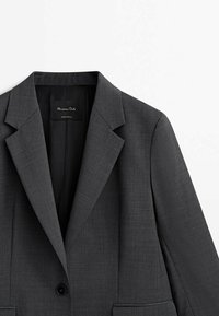 Blazer grigio scuro con sottogola a punta, chiusura a un bottone e tessuto texturizzato. Etichetta visibile nell'area del colletto. Design semplice, senza tasche.