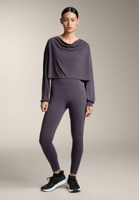 Ljubičasti skraćeni dugi rukav majica s drapiranim dekolteom u kombinaciji s prilagodljivim ljubičastim tajicama i crnim sportskim cipelama. Glatka tekstura.