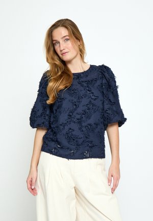 Žena s dlhými blond vlasmi má na sebe námornícky modrý textúrovaný top s nadýchanými rukávmi a krémové široké nohavice, stojaca pred jednoduchým bielym pozadím.