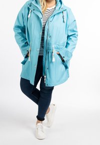 Modrá voděodolná bunda s kapucí, šňůrkami a třásněmi. Kombinována s tmavými džínami a bílými teniskami. Pod ní pruhované tričko.
